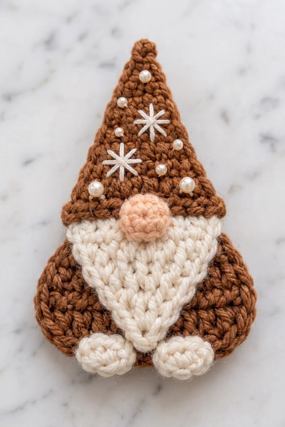 A crocheted gingerbread gnome with brown hat, cream beard, and pearl bead decorations on marble background.