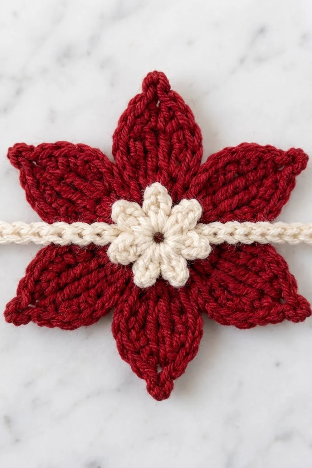 A crocheted red poinsettia flower with cream center and twisted cord, displayed on marble.