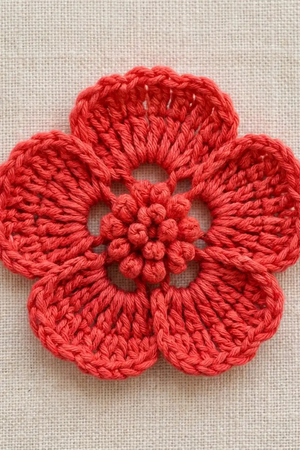 A bright red crocheted flower with five petals and textured center detail rests on a neutral beige fabric background.