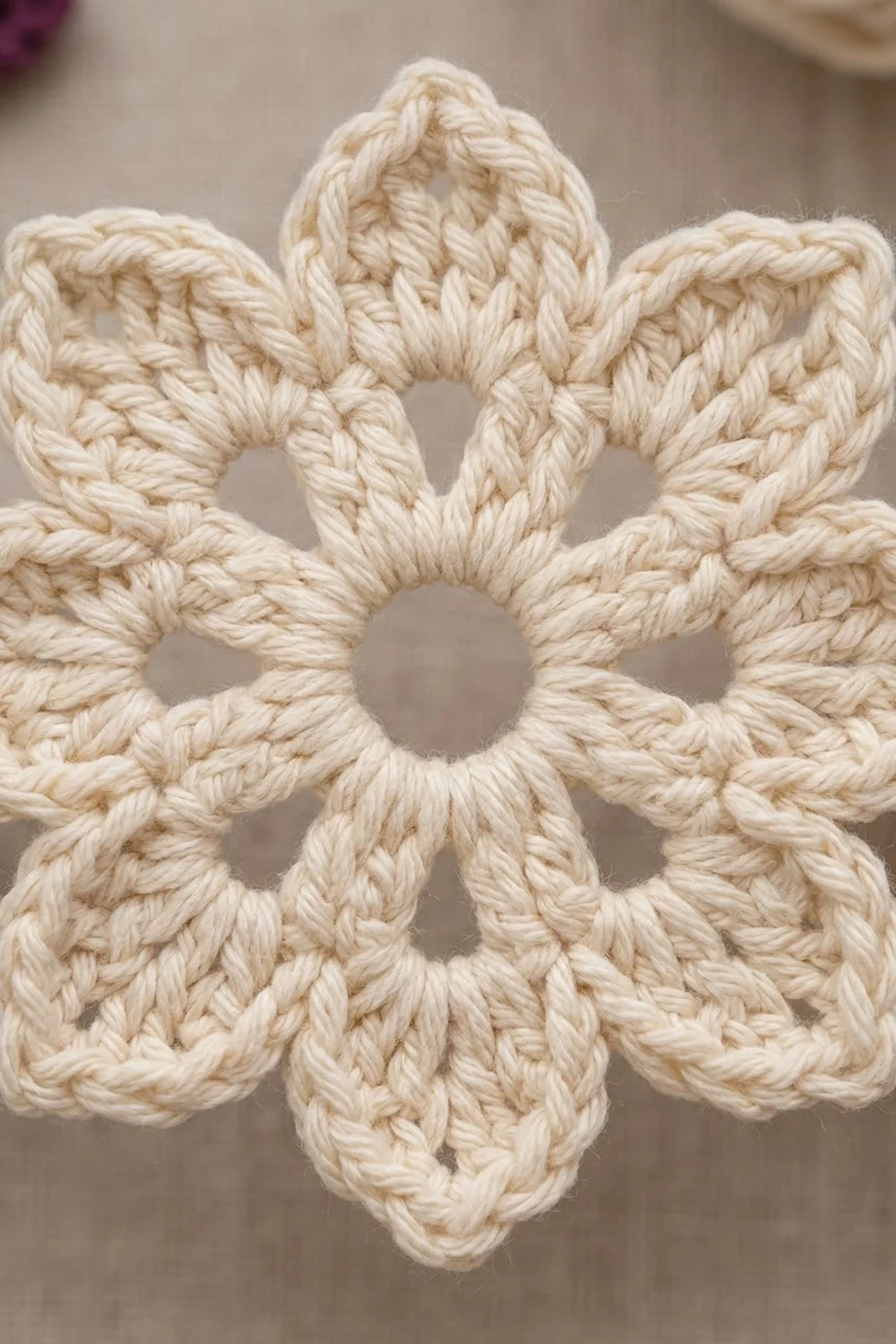 A cream-colored crocheted flower with six petals and an open center displayed against a soft beige background.