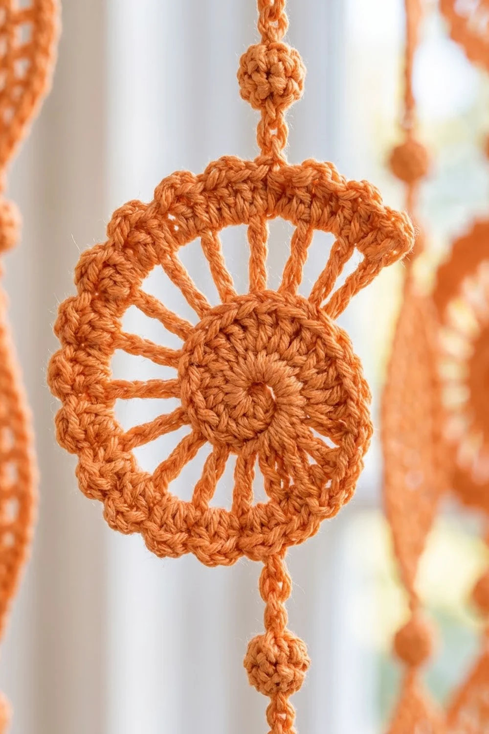Orange crocheted ornament with sun motif hanging by cord against blurred window background.