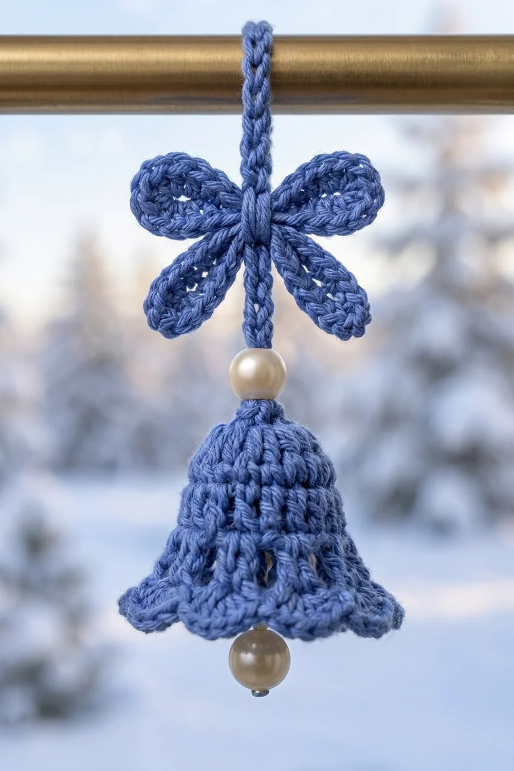 Blue crocheted bell ornament with pearl beads hanging from a brass rod against snowy background.
