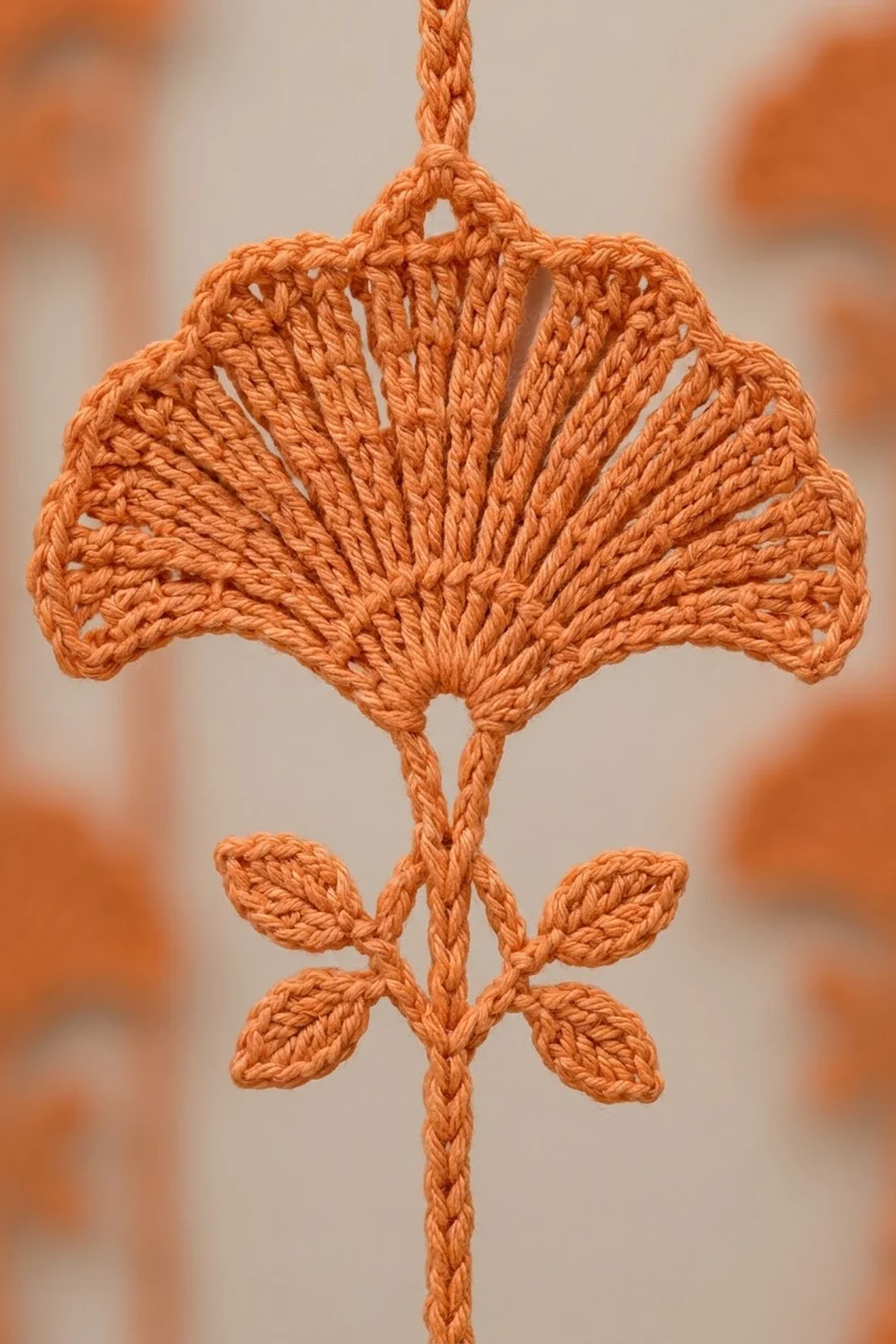 An orange crocheted ginkgo leaf ornament with small leaves at the base, hanging against a soft blurred background.
