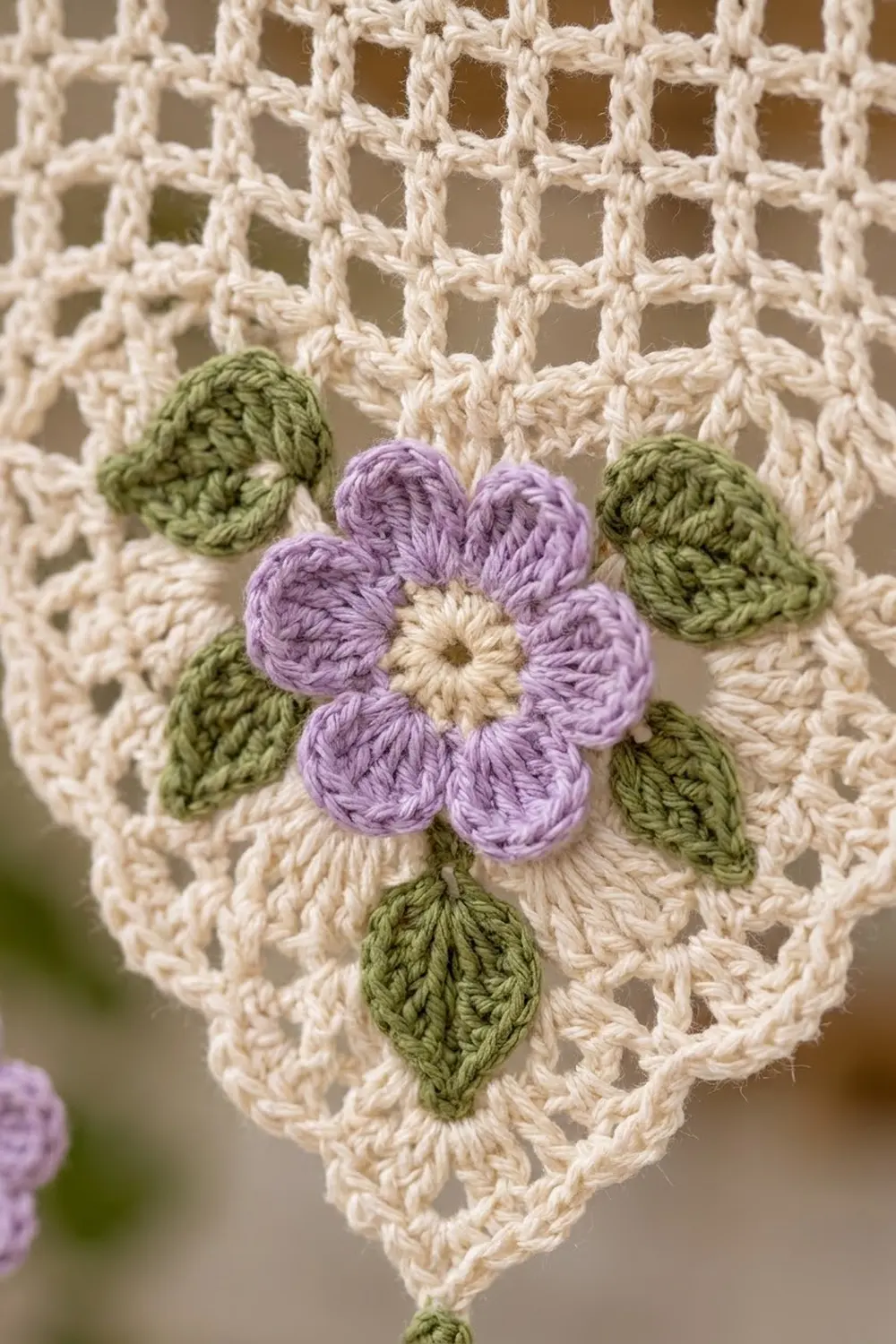 A crocheted purple flower with cream center and green leaves attached to a cream lace macramé panel.