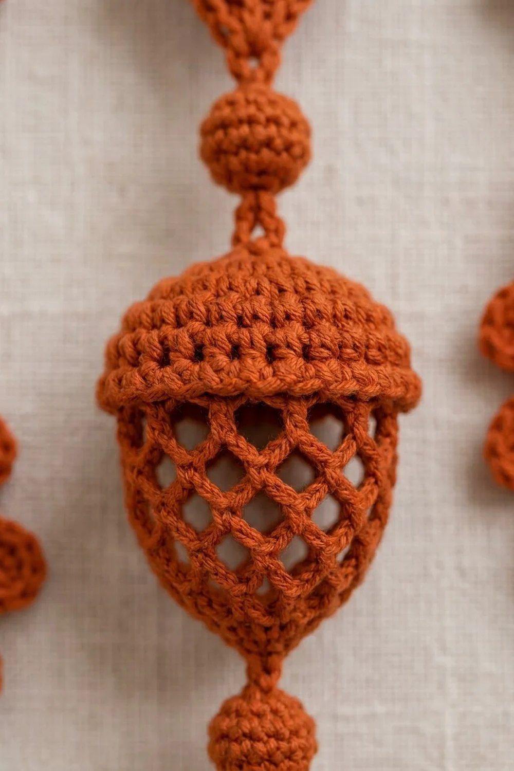 Close-up of an orange crocheted acorn ornament with lattice pattern hanging against a white linen background.