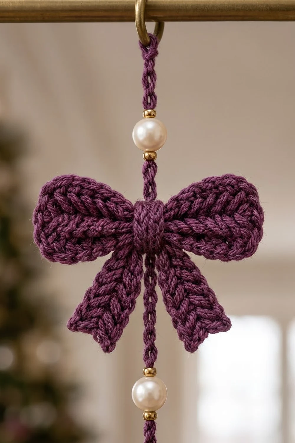 A purple crocheted bow ornament with pearl beads hanging from a brass ring against a blurred background.