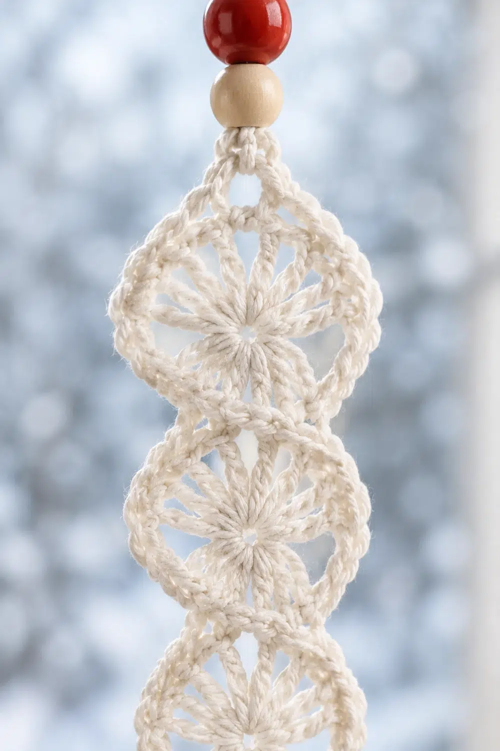 A cream-colored macramé ornament with diamond patterns topped by a natural wood bead and red ball.