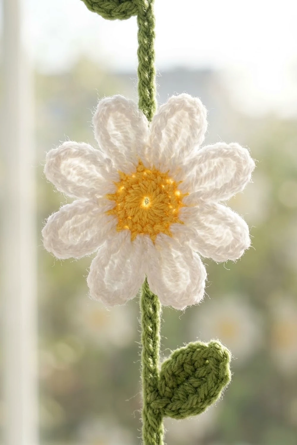 A crocheted white daisy with yellow center hanging from a green braided stem against a blurred outdoor background.