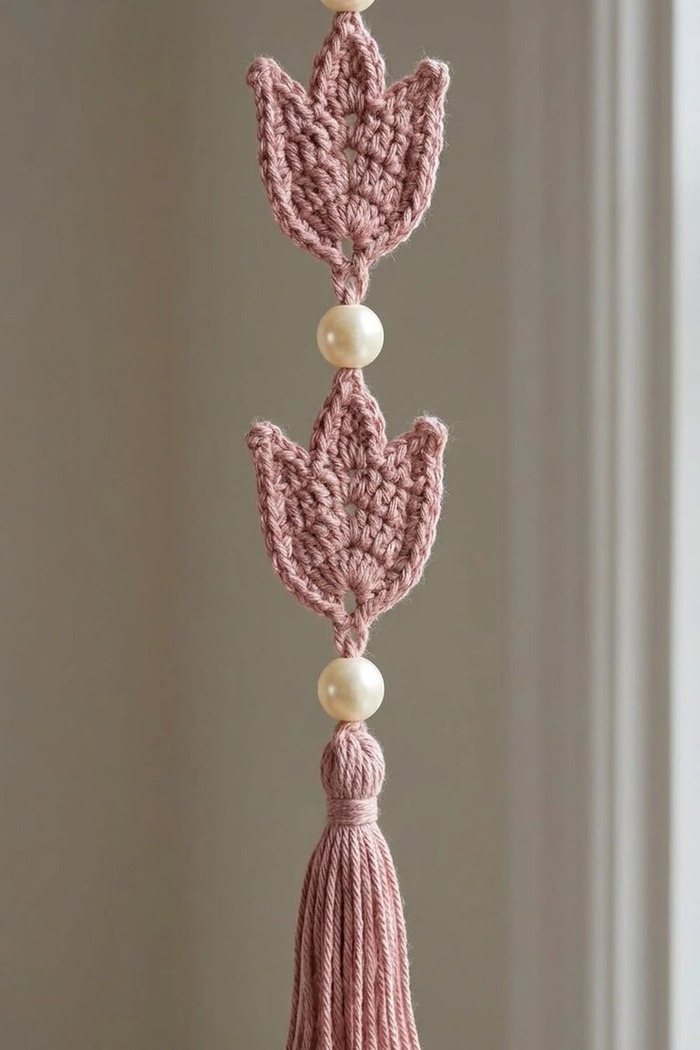 A crocheted tulip garland in dusty rose with cream beads strung vertically against a neutral wall.