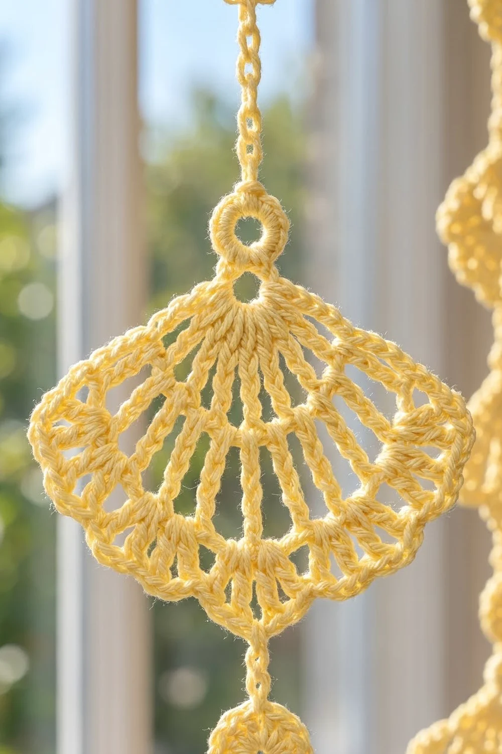 A golden yellow crocheted diamond-shaped ornament hanging by a chain in front of a window.