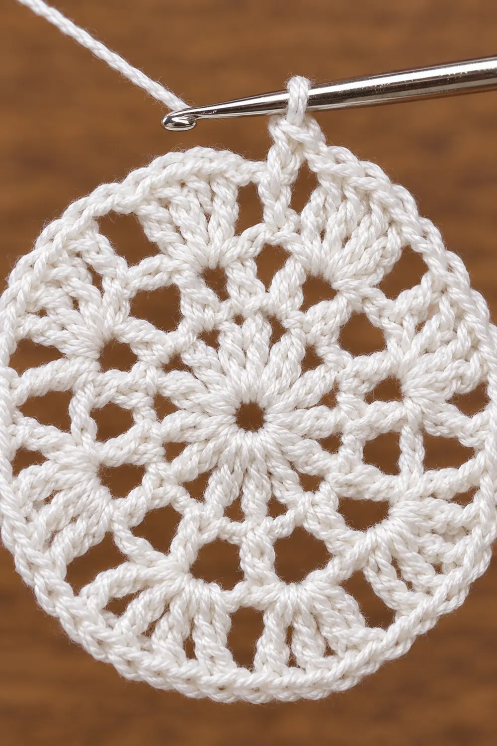 White crocheted snowflake ornament hanging from a silver crochet hook against a brown background.