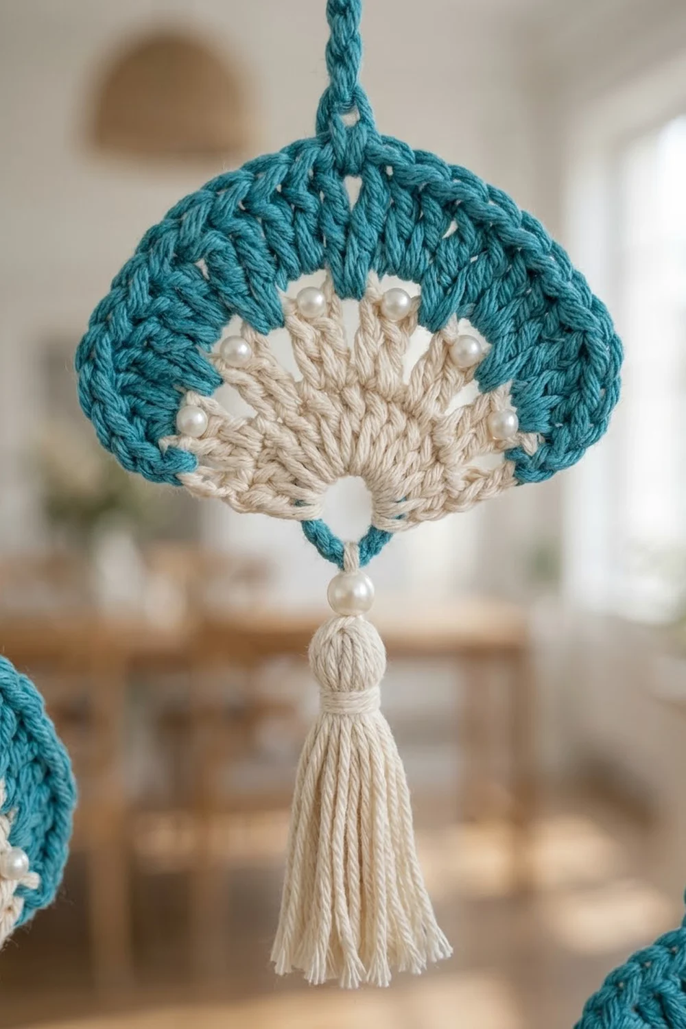 A handmade macramé ornament with teal top, cream center, pearl beads, and tassel hanging indoors.