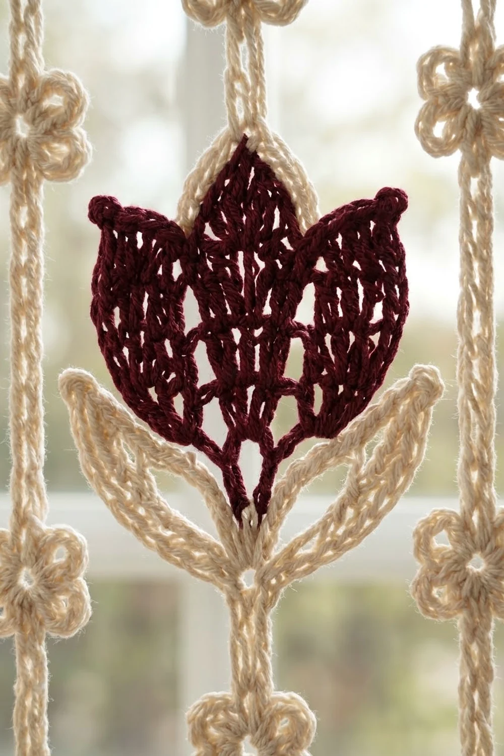 Crocheted burgundy heart ornament with openwork pattern suspended by tan rope against a bright window.