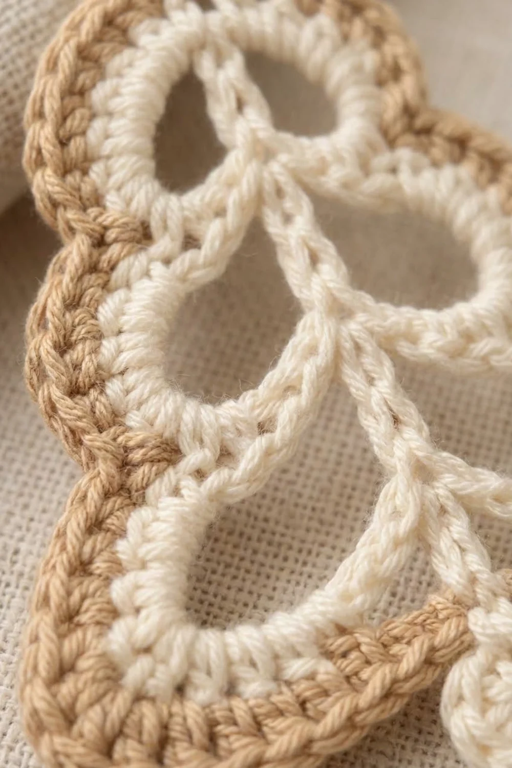 Close-up of crocheted chain loops in cream and tan yarn on a neutral textured fabric background.