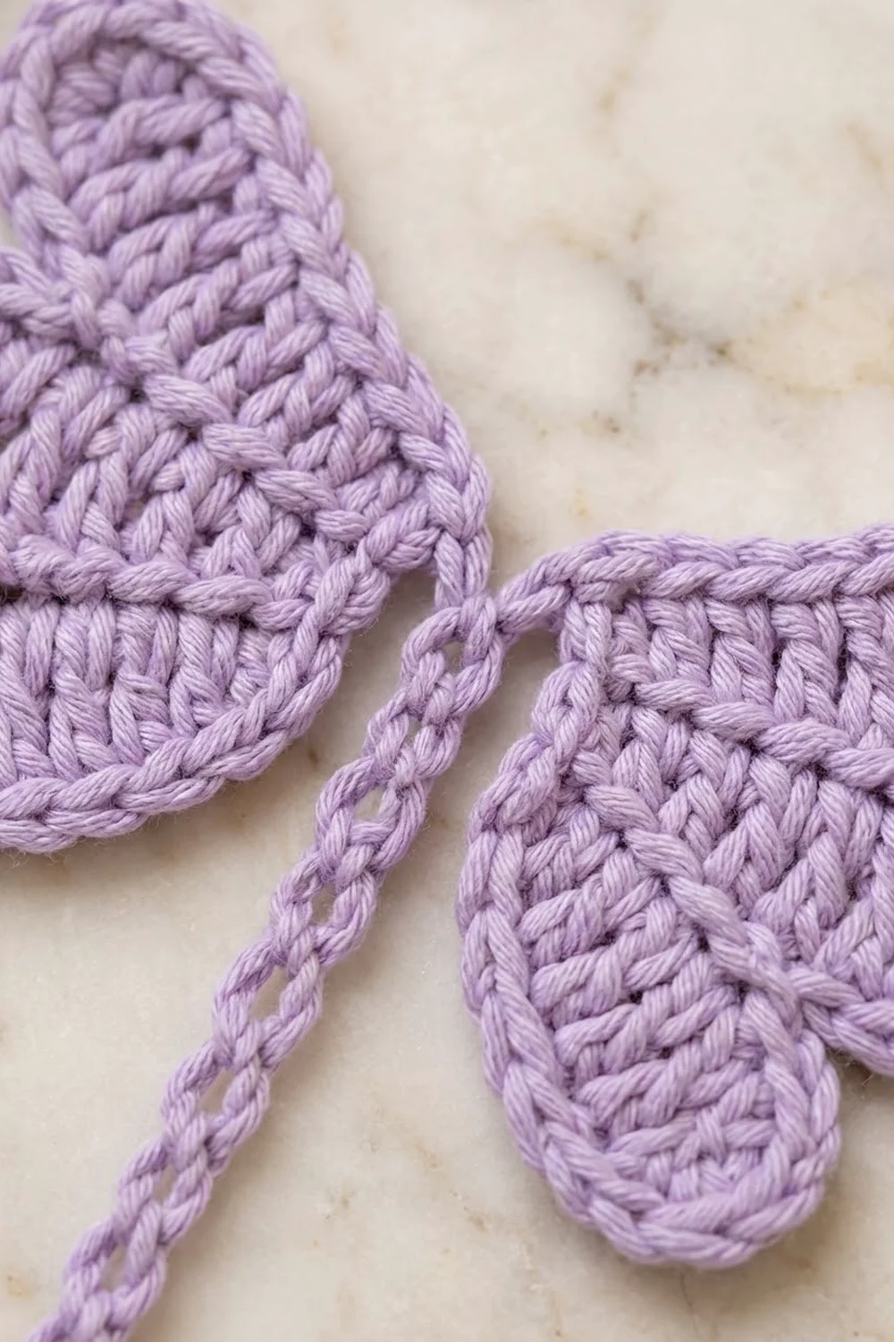 Close-up of lavender crocheted mittens with braided cord straps on a cream marble surface.