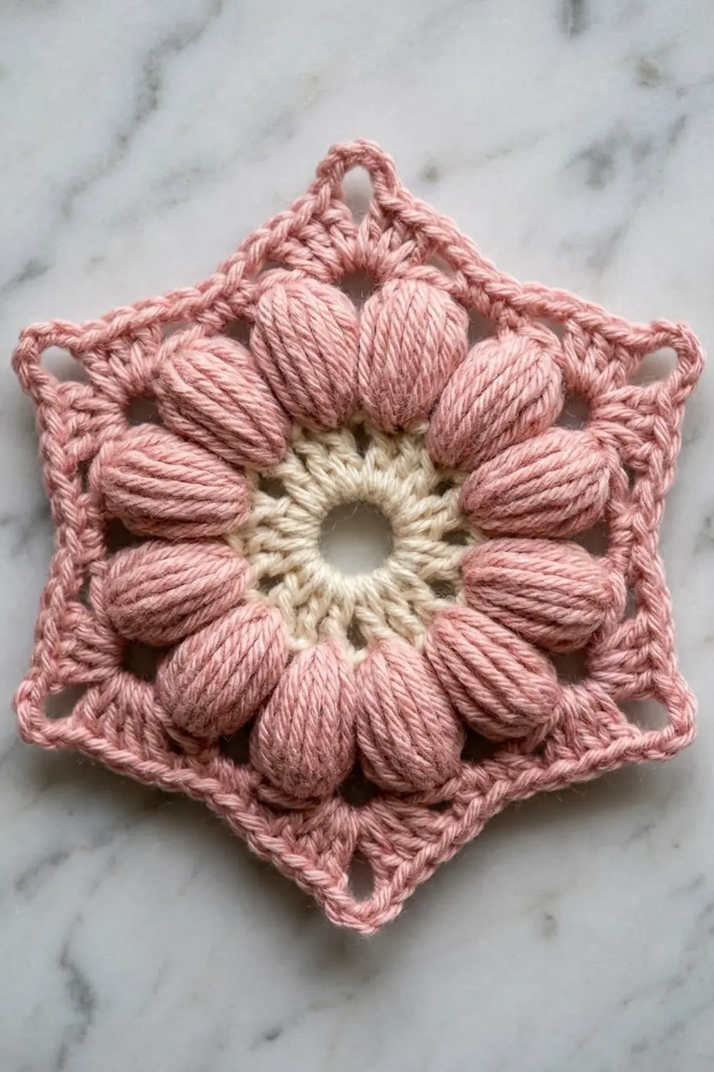 A crocheted pink and cream flower ornament with textured petals displayed on white marble background.