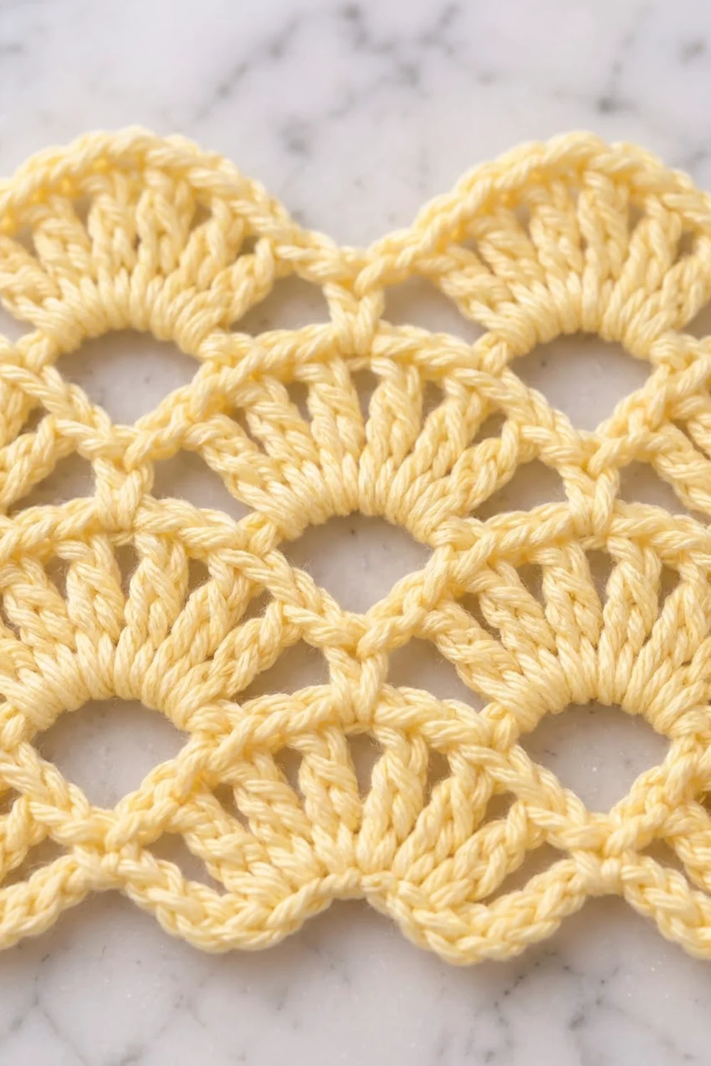 Close-up of pale yellow crochet lace with interlocking loops on a marble surface.