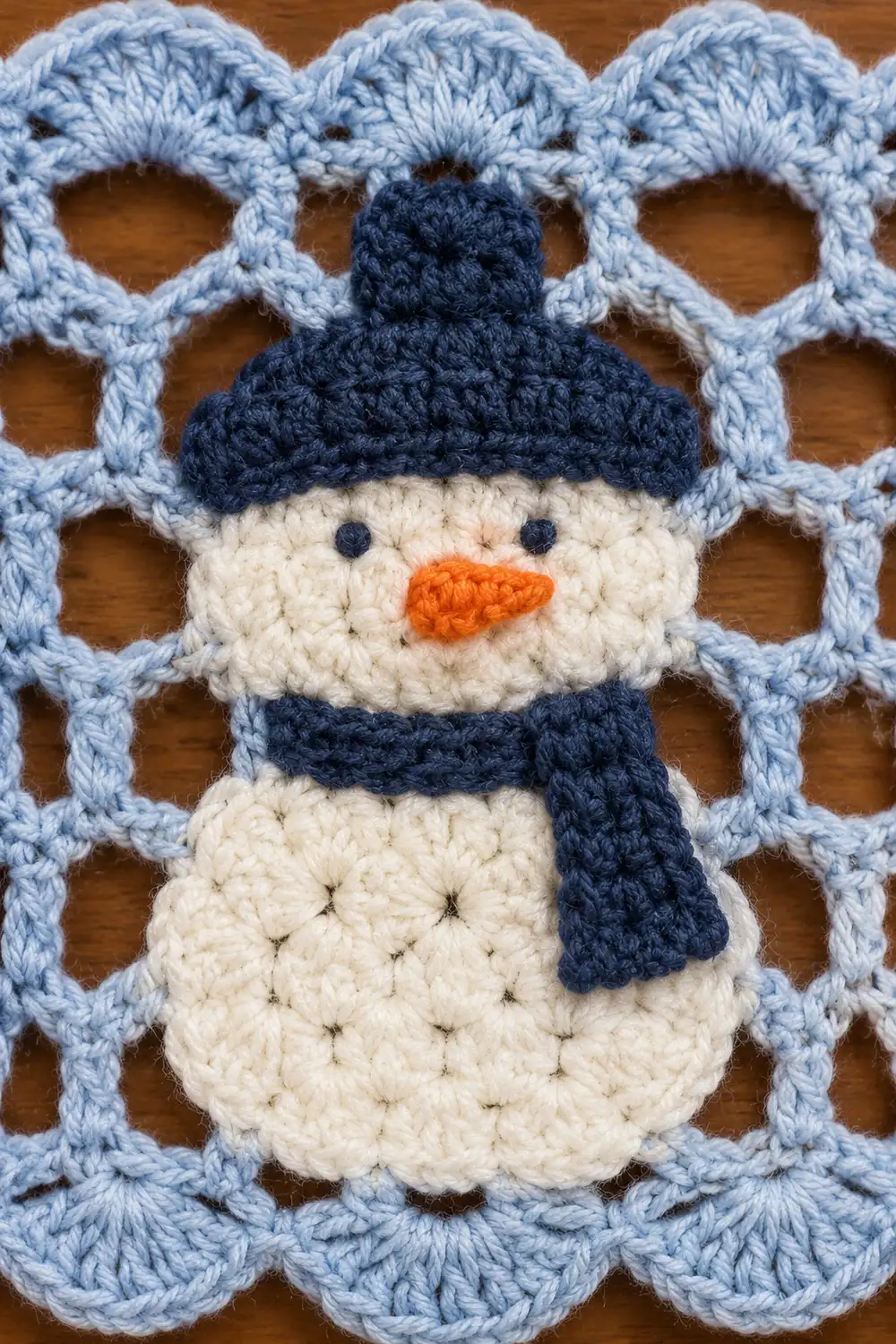 A crocheted snowman ornament with navy hat and scarf on a light blue lattice crochet background.