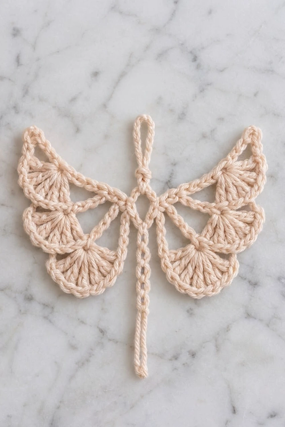 A crocheted butterfly ornament in cream-colored yarn with a loop, displayed on marble surface.