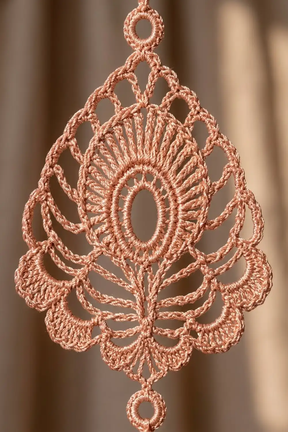 A delicate peach-colored crocheted lace pendant with intricate teardrop patterns hangs against a soft blurred background.