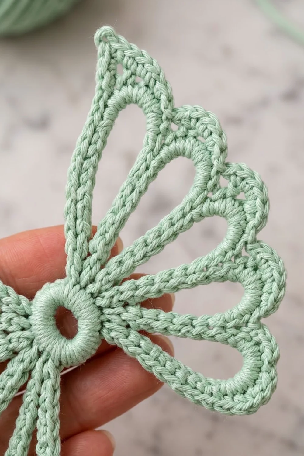 A delicate pale green crocheted or macramé bow held in someone's hand against a blurred neutral background.
