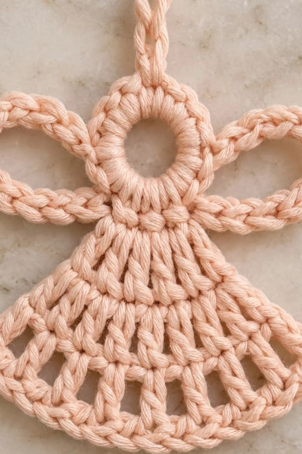 A peach-colored macramé angel ornament with twisted rope details displayed against a marble background.