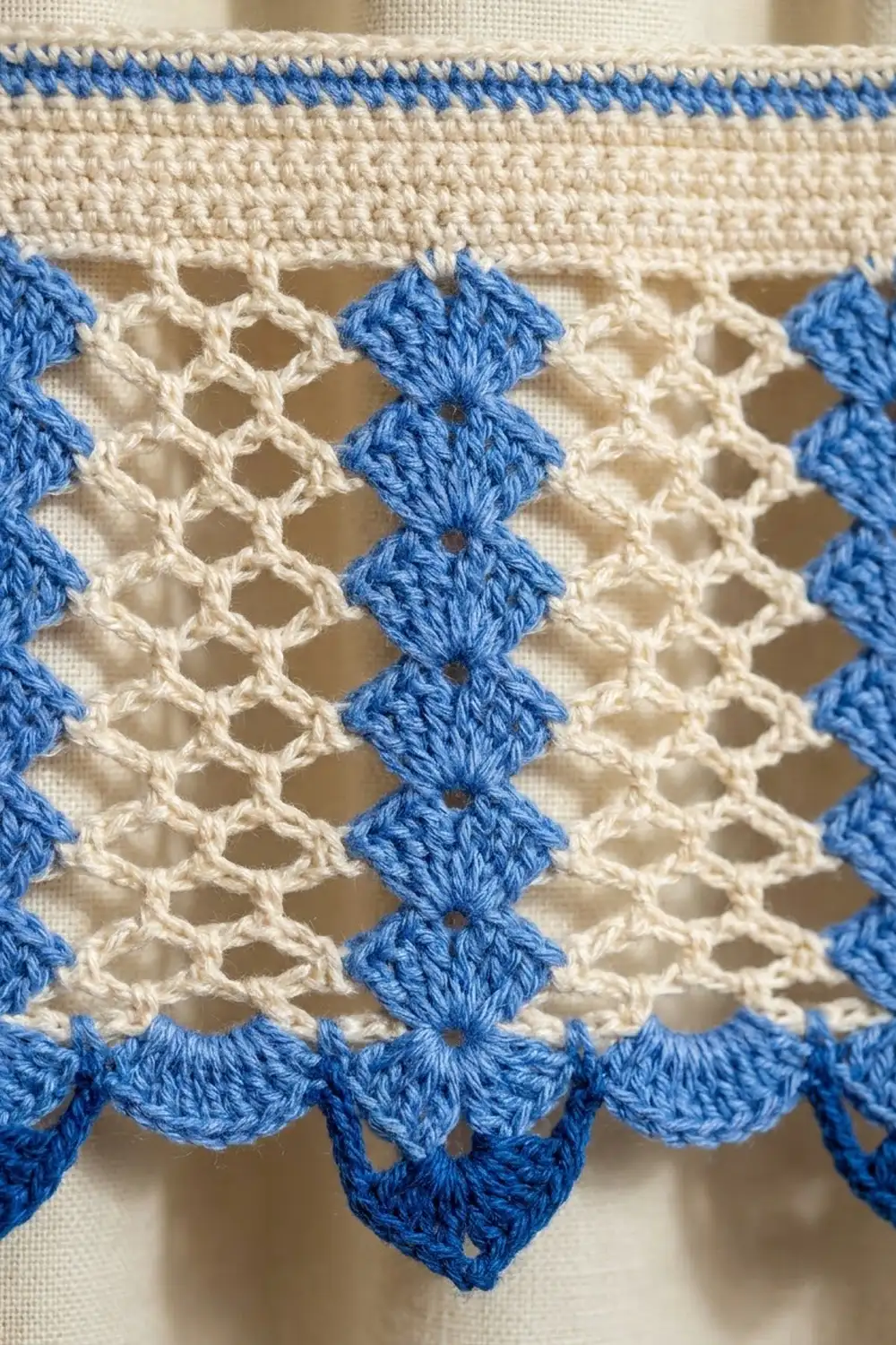 Close-up of cream and blue crocheted trim with diamond motifs and scalloped edging on fabric background.