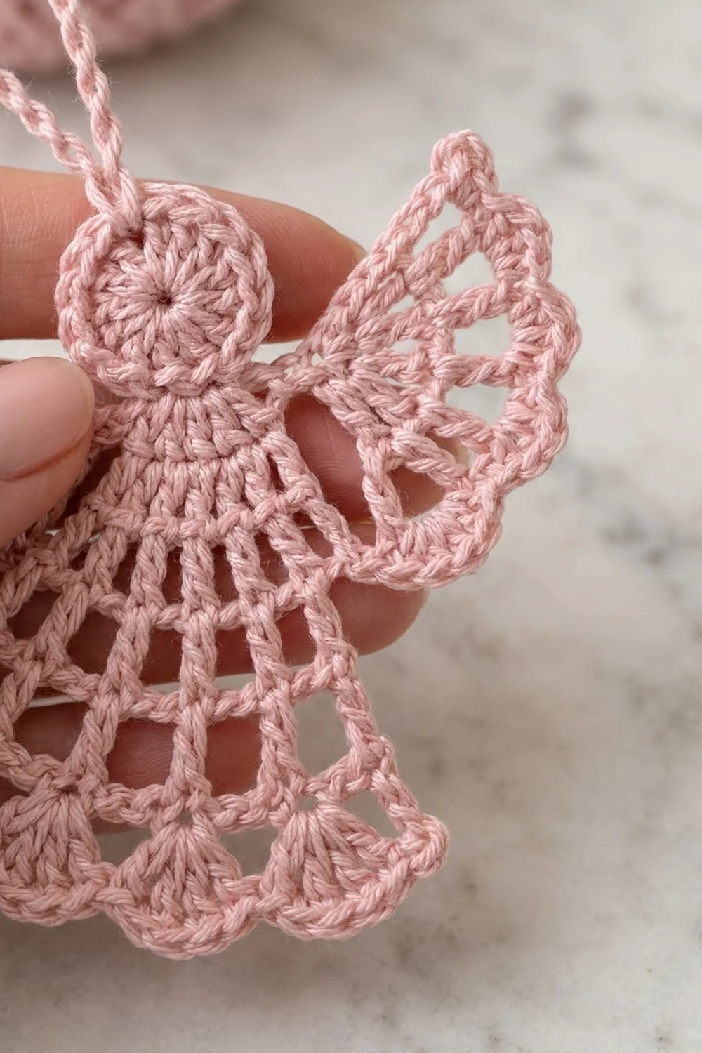 A delicate pink crocheted angel ornament held in someone's hand, displaying detailed lacework wings and body.