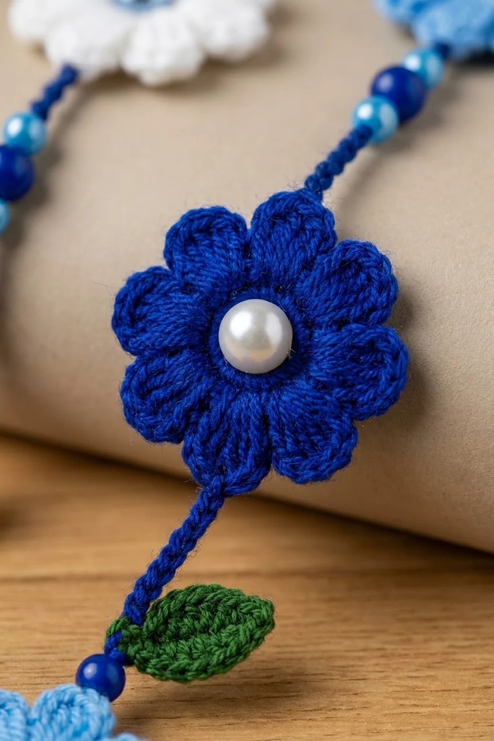 A crocheted blue flower with a white pearl center and green leaf on a beaded string, displayed on wood.