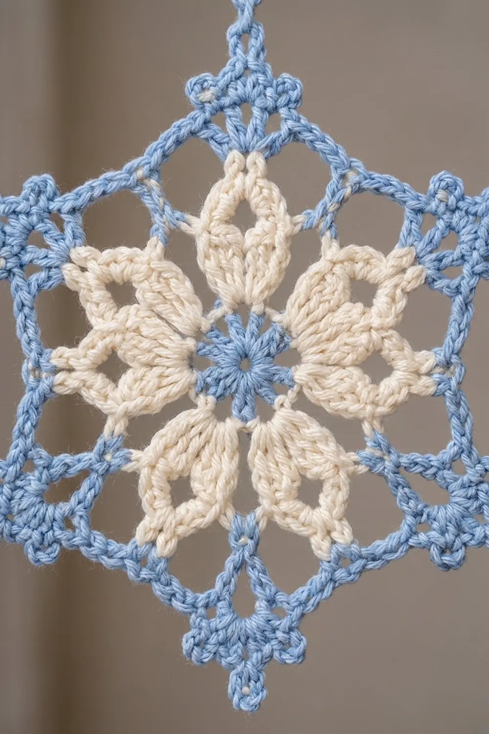 A crocheted snowflake ornament with blue outer edges and cream-colored center petals hanging against a blurred beige background.