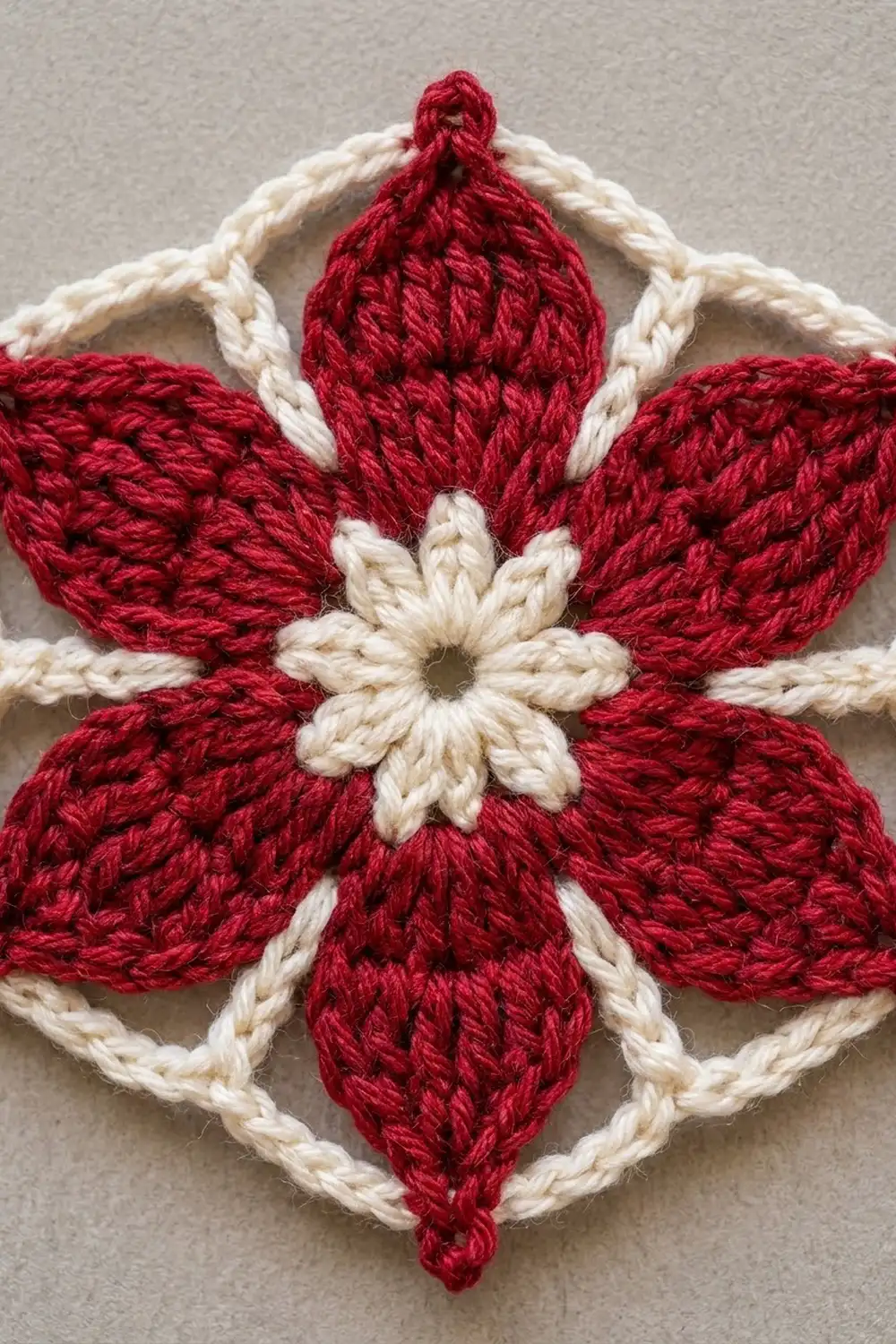 Crocheted red and cream flower ornament with textured petals and a cream center on a gray background.