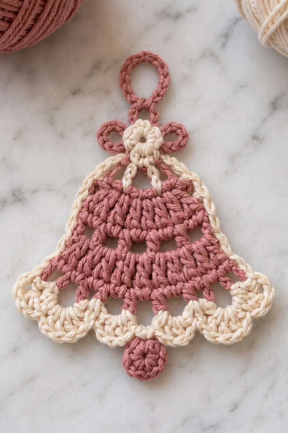 A crocheted bell ornament in dusty rose and cream yarn displayed on a marble surface.
