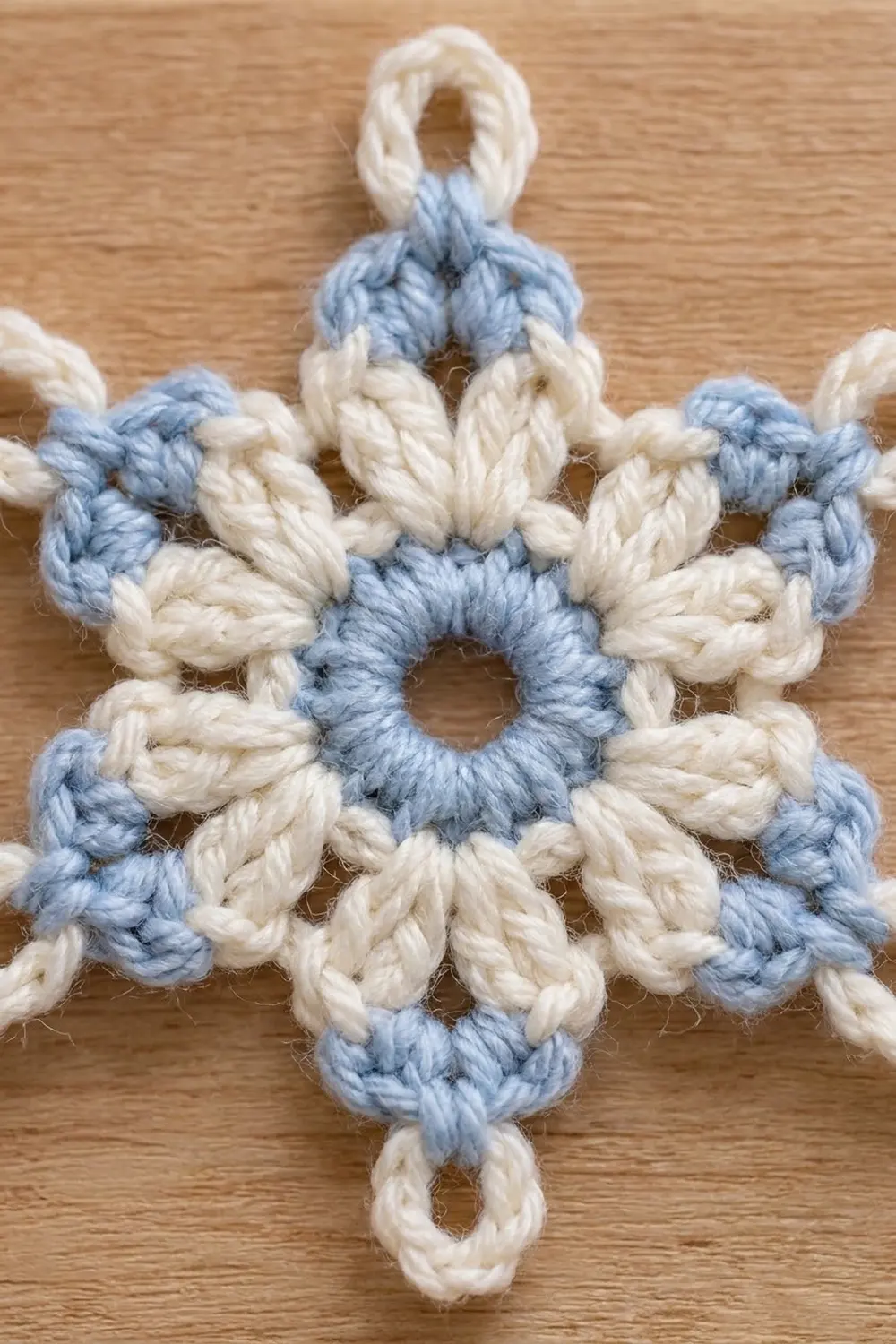 A crocheted snowflake ornament in blue and cream yarn with loops at top and bottom, displayed on wood.