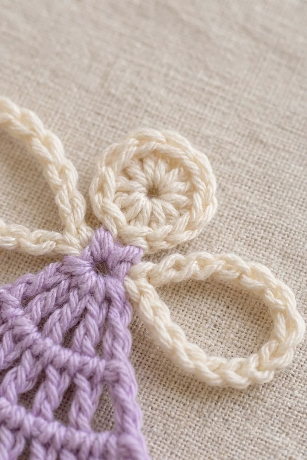 Crocheted flower appliqués in cream and lavender purple yarn arranged on natural linen fabric background.