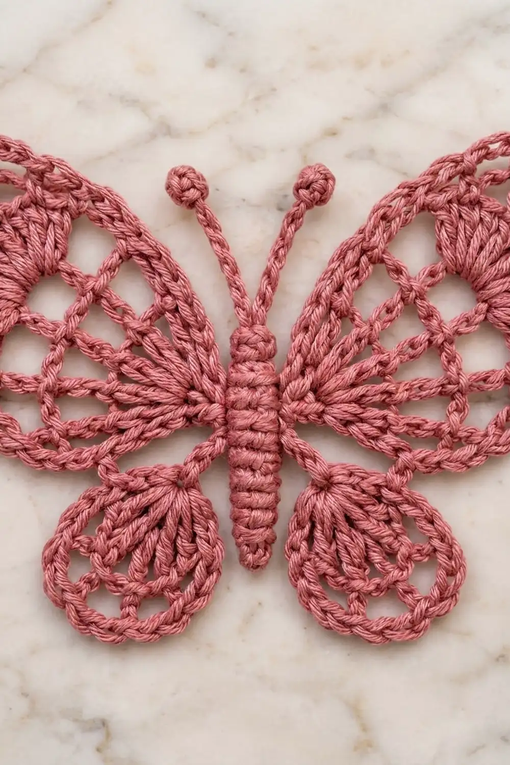 A delicate pink crocheted butterfly appliqué with intricate lace wings displayed on a marble surface.