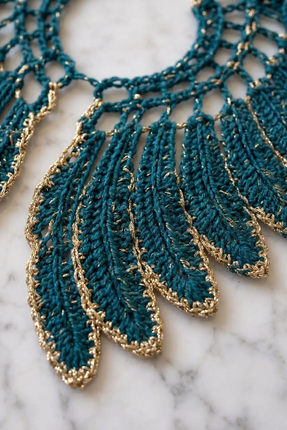 Teal and gold crocheted lace collar with dangling leaf-shaped fringe displayed on marble surface.