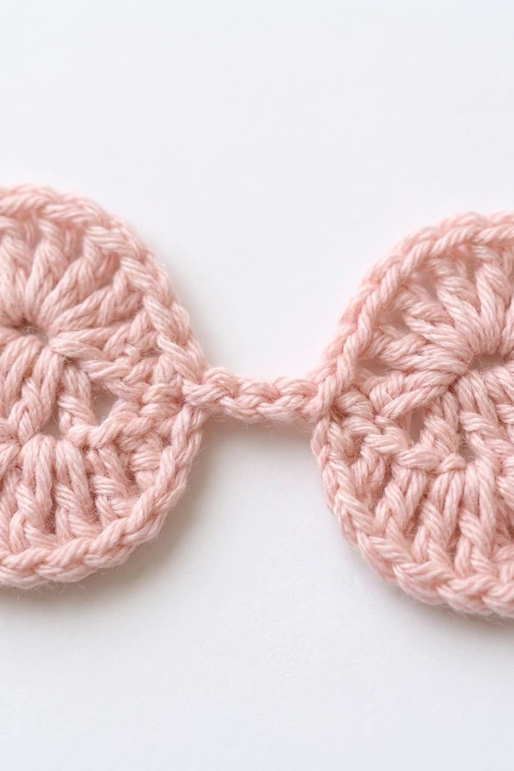 Close-up of two pink crocheted or knitted bow shapes connected by yarn against a white background.
