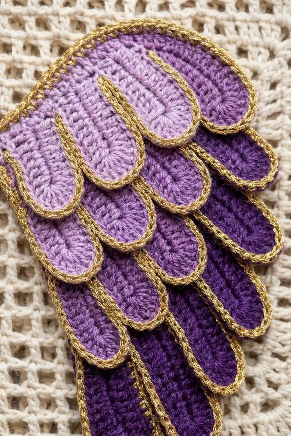 Crocheted purple flower with layered petals outlined in gold yarn displayed on a cream-colored textured background.