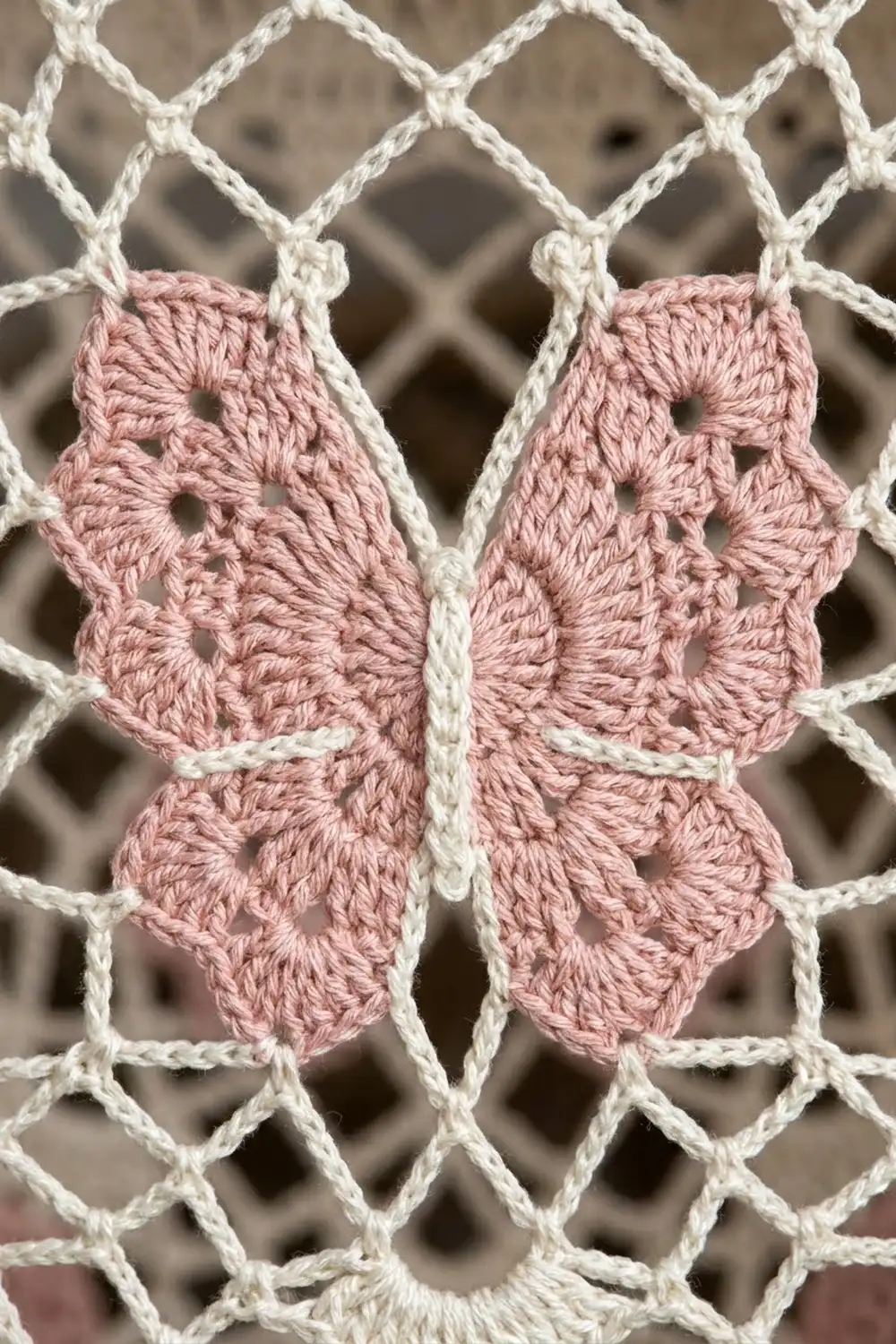 A delicate pink crocheted butterfly appliqué attached to cream-colored macramé netting with intricate knots.