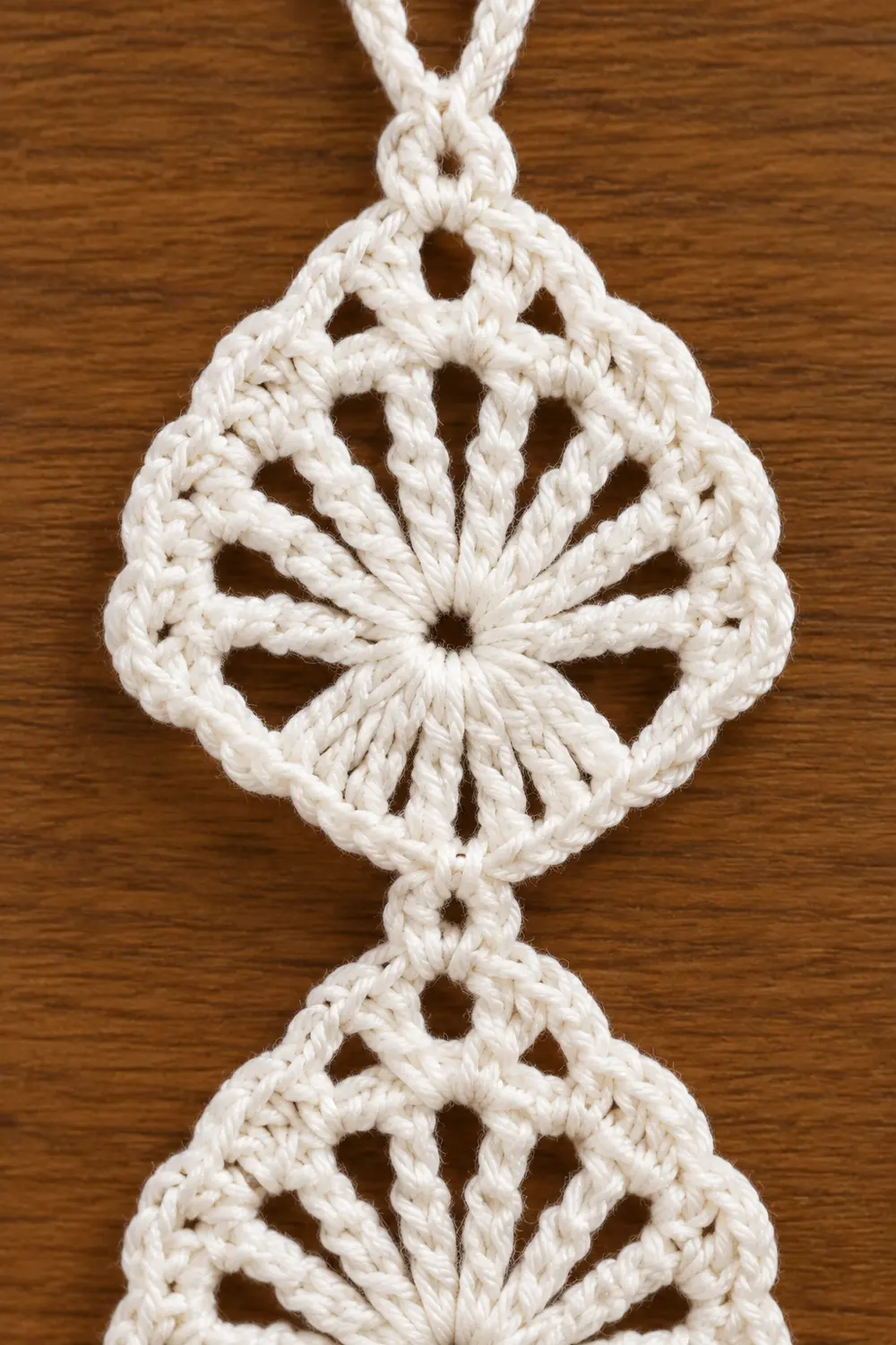 Cream-colored macramé ornament with two connected diamond-shaped medallions displayed on a wooden surface.
