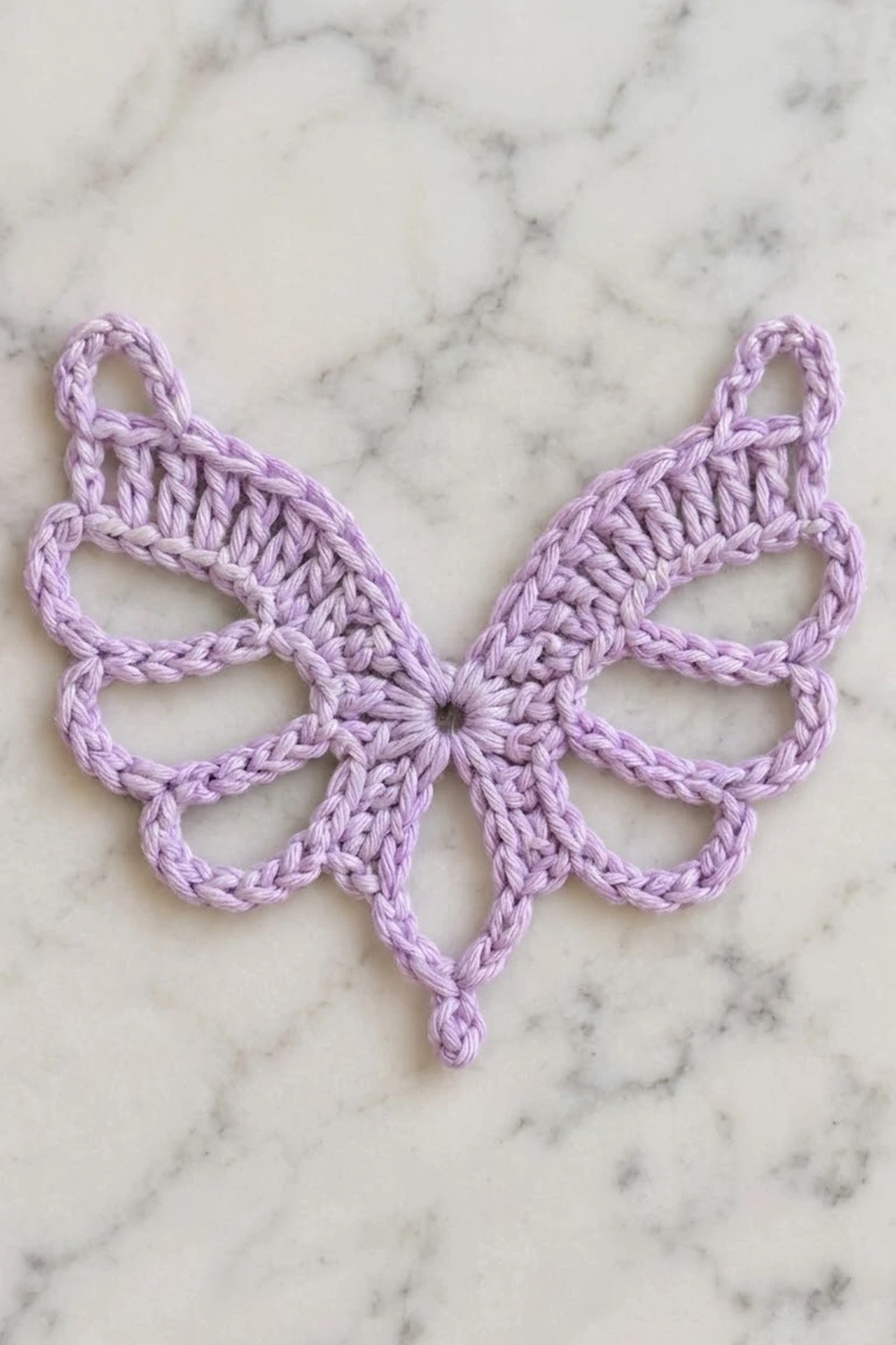 A lavender crocheted butterfly motif with looped petals displayed on a white marble surface.