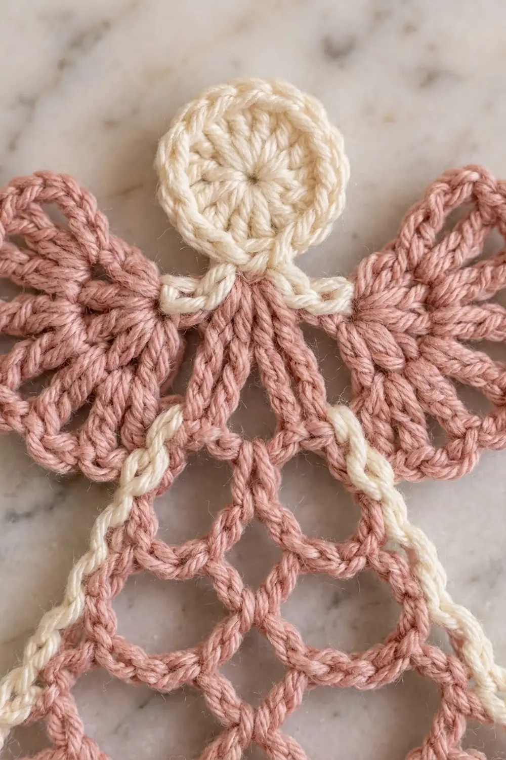 Crocheted angel ornament with cream head and body, dusty rose wings, displayed on marble surface.