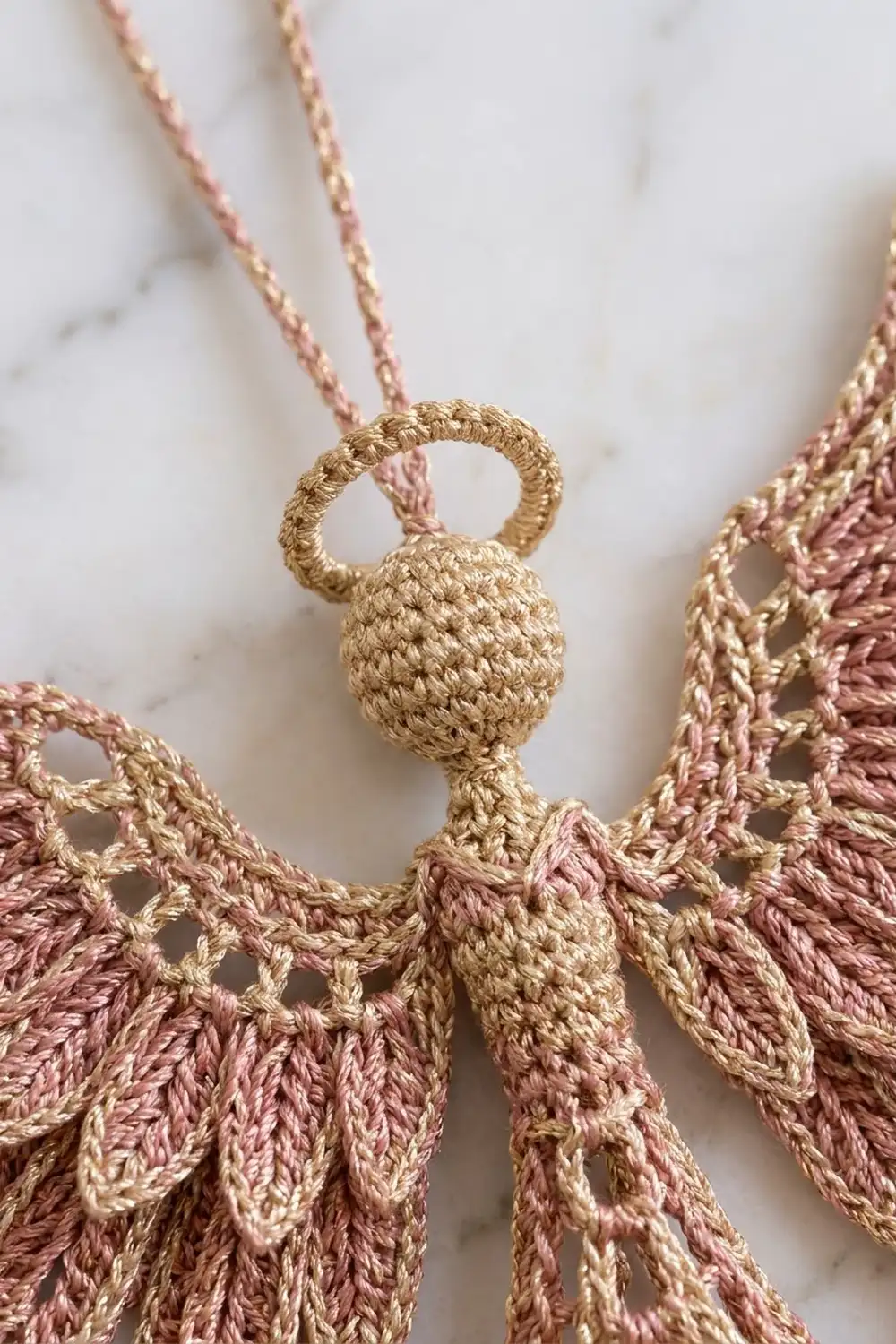 Close-up of a crocheted angel ornament with mauve and cream wings and a beaded head against marble background.