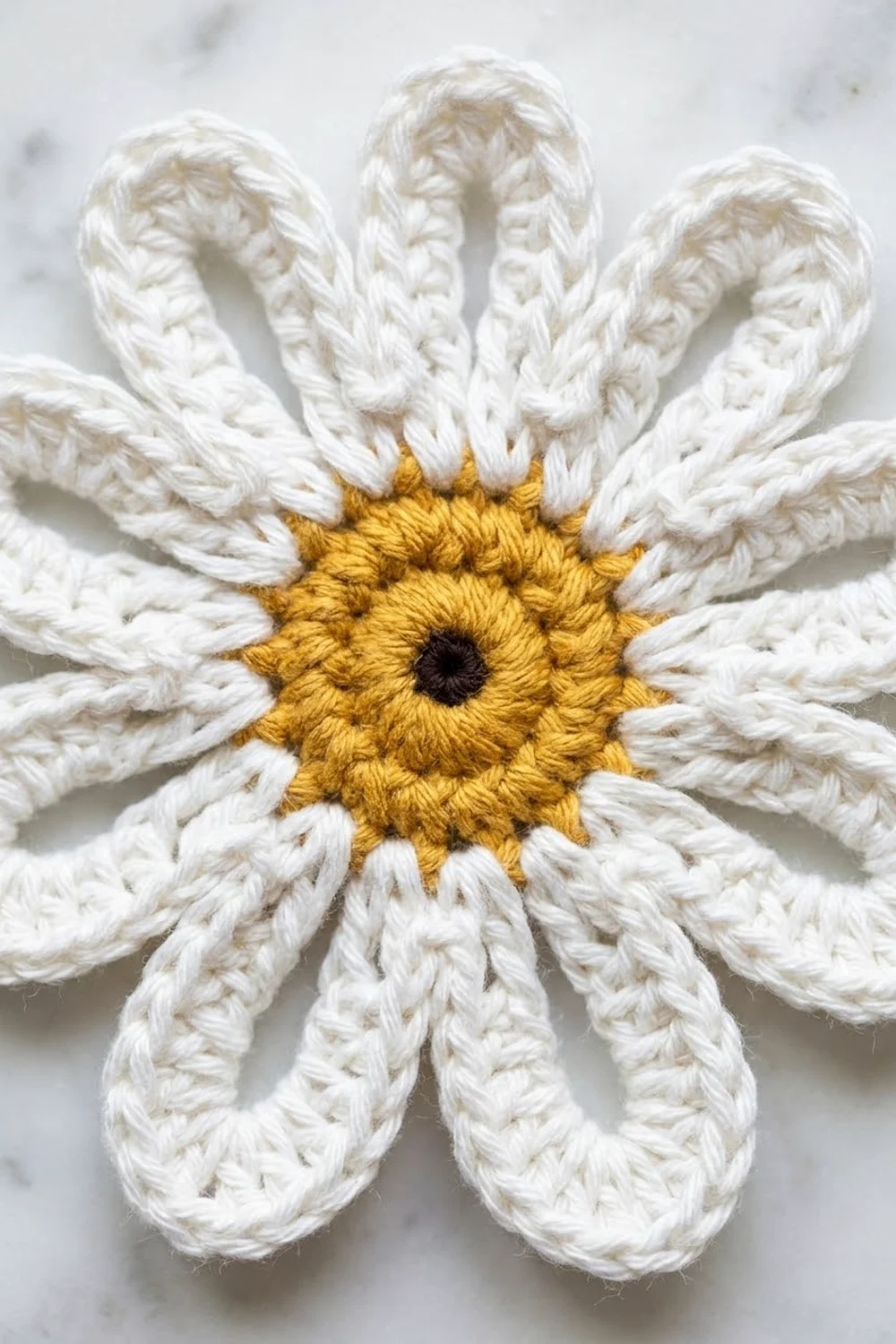 A crocheted daisy with white petals and a golden yellow center with black detail on a white background.