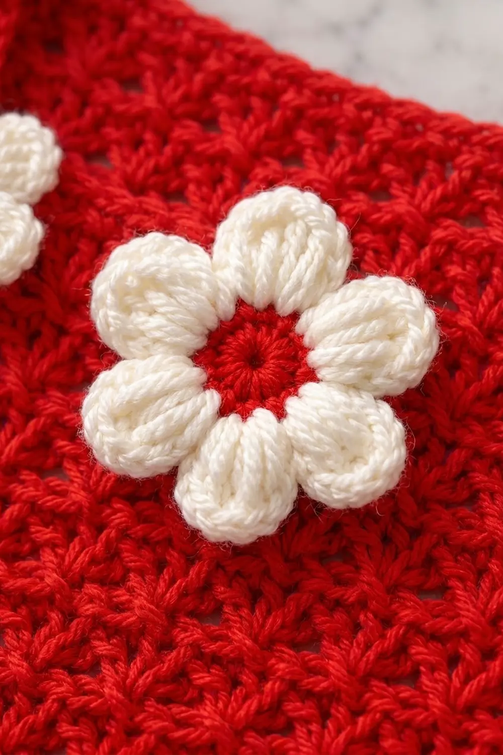 A white crocheted flower with six petals adorns a red textured crocheted fabric background.