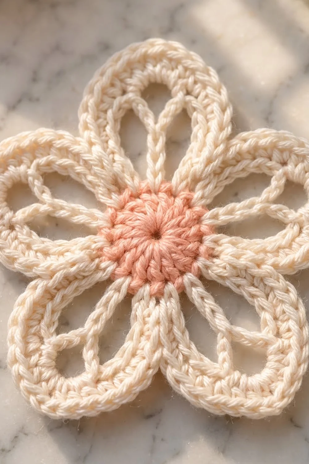 Crocheted flower ornament with cream petals and coral pink center on marble surface.