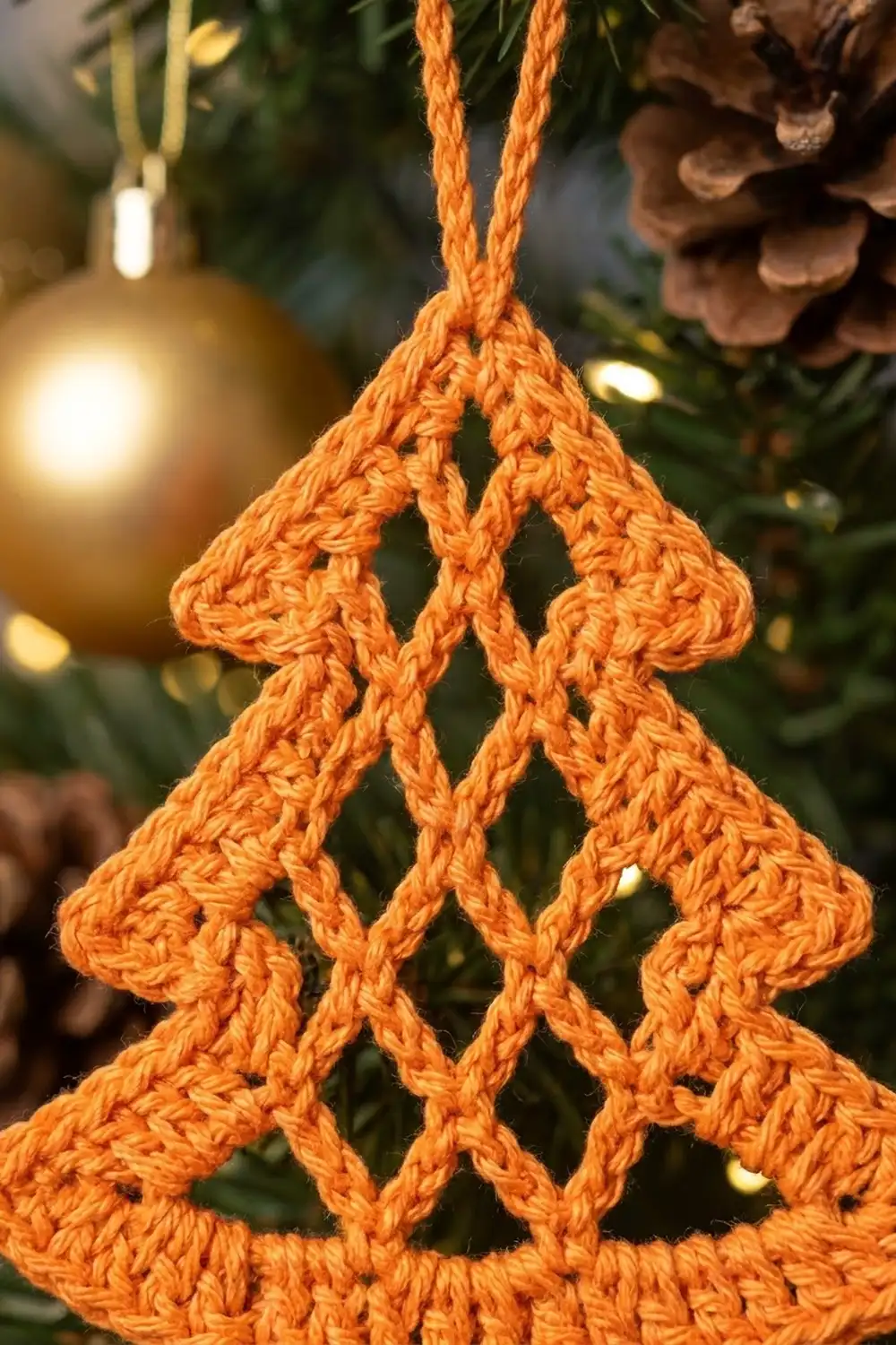 A crocheted orange Christmas tree ornament hangs among evergreen branches and gold baubles.