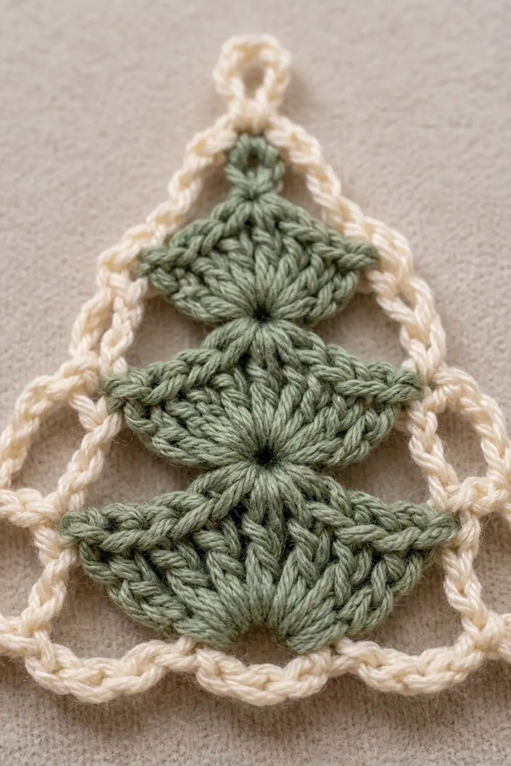 Crocheted Christmas tree ornament with sage green body and cream-colored scalloped border on neutral background.