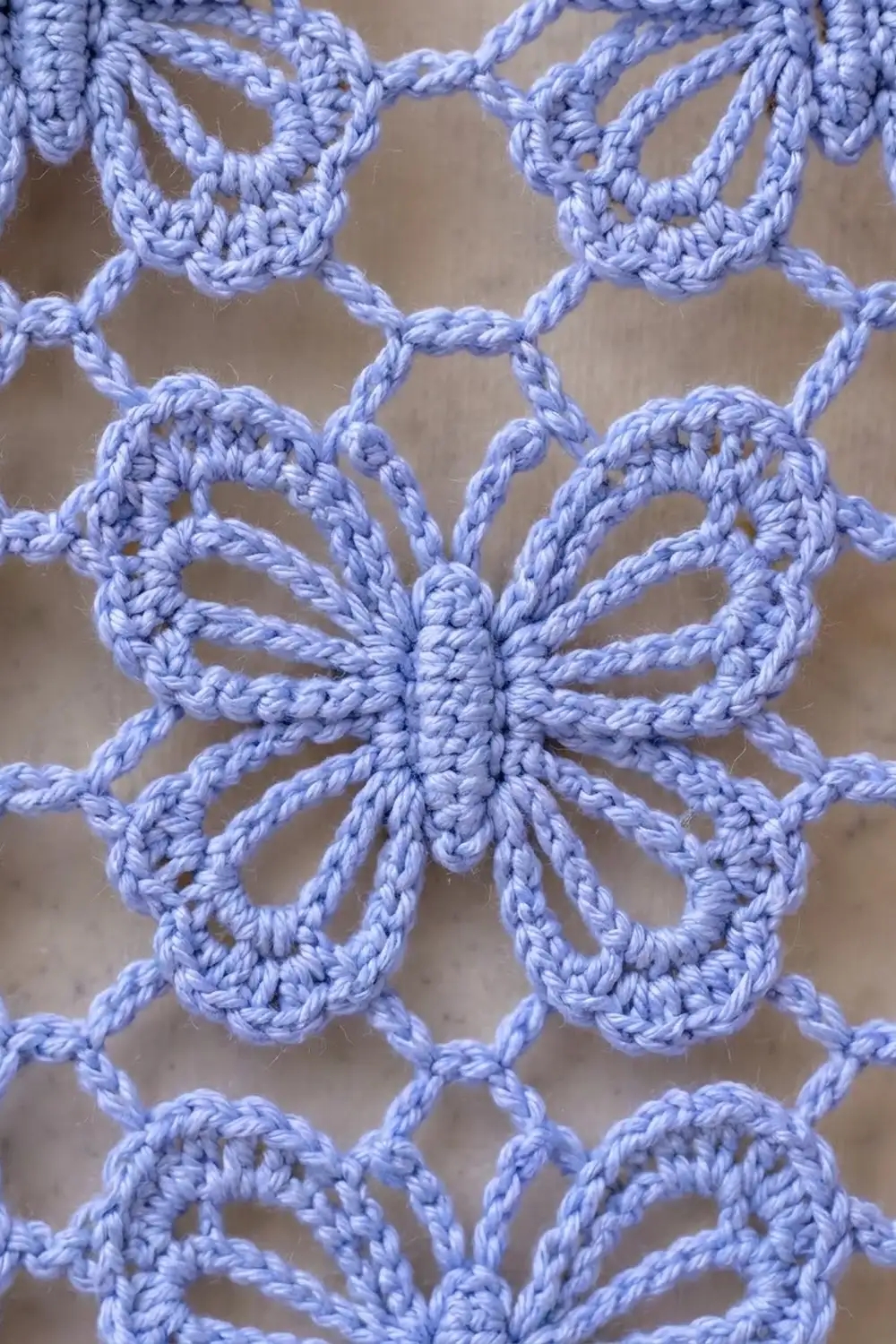 Close-up of delicate periwinkle blue crocheted lace with repeating butterfly and floral motifs against a neutral beige background.