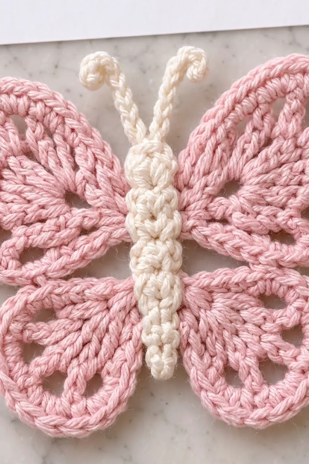A crocheted butterfly with pink wings and cream-colored body and antennae rests on a marble surface.